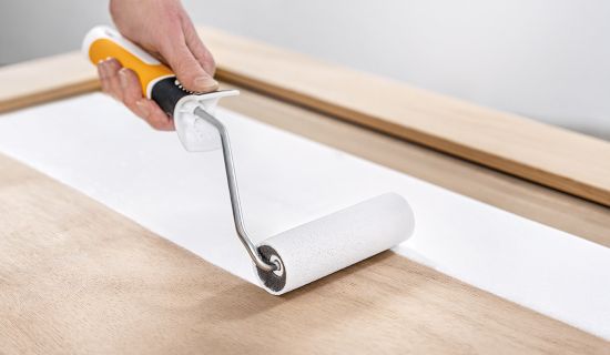 Eine Hand mit einem gelb-weißen Griff führt eine kleine Schaumstoff-Lackrolle über eine glatte, helle Holztür. Die Rolle hinterlässt eine saubere, deckende Bahn weißer Farbe auf dem unbehandelten Holz.