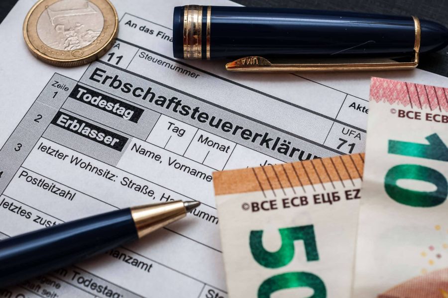 Nahaufnahme eines offiziellen Formulars zur Erbschaftsteuererklärung, auf dem Schreibgeräte, eine Euro-Münze und Euro-Geldscheine liegen.