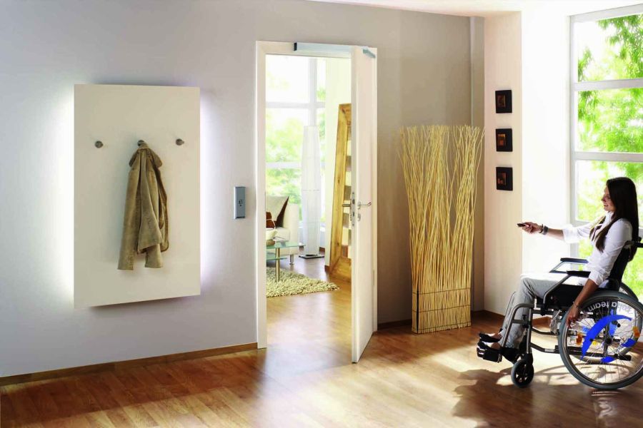 Eine Frau im Rollstuhl sitzt in einem hellen, modernen Raum und hält eine Fernbedienung in der Hand. Im Hintergrund ist eine offene Tür zu sehen, die auf einen weiteren Bereich des Raums führt, und an der Wand hängt ein Mantel an einem weißen Garderobenpa