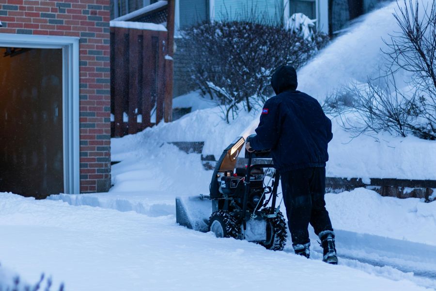 Eine Person in dunkler Winterkleidung schiebt eine motorisierte Schneefräse über einen verschneiten Gehweg oder eine Einfahrt. Hoher Schnee türmt sich an den Rändern auf, während die Maschine den Schnee zur Seite wirft.