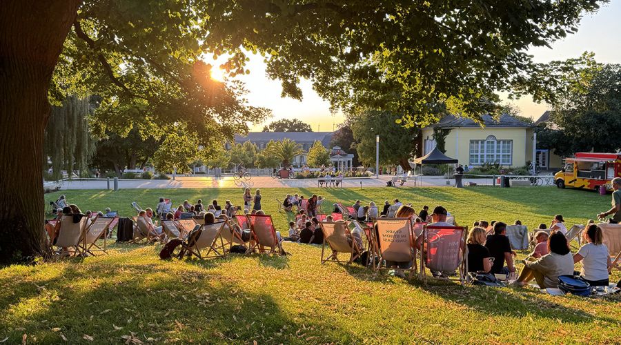 Auf einer Wiese im Abendlicht sitzen viele Menschen gemütlich in Liegestühlen