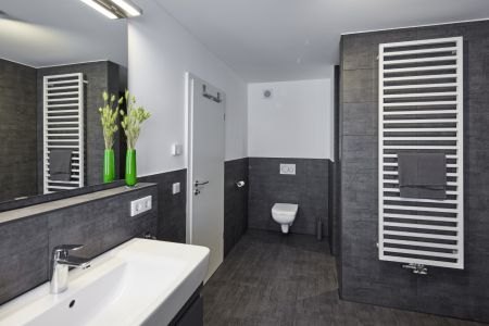 Ein modernes Badezimmer mit dunkelgrauen Fliesen und weißen Wänden. Zu sehen sind ein Waschbecken, eine Toilette, ein großer Spiegel sowie ein weißer Handtuchheizkörper. Oben an der Wand ist ein kleines, weißes Lüftungsgitter erkennbar.