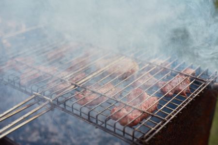 Eine Nahaufnahme von Fleischstücken, die in einem Grillkorb über glühender Kohle garen. Dichter, weißer Rauch steigt vom Grill auf und hüllt das Grillgut ein.