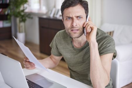 Ein Mann sitzt an seinem Schreibtisch vor einem Laptop und telefoniert mit dem Smartphone am Ohr. In der anderen Hand hält er ein Dokument und blickt mit einem skeptischen, leicht irritierten Gesichtsausdruck in den Raum.