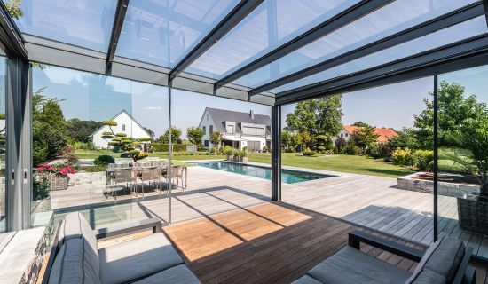 Ein lichtdurchfluteter Wintergarten mit Glasdach und Schiebewänden bietet einen Panoramablick auf einen Pool und einen gepflegten Garten mit modernen Häusern. Im Innenraum stehen ein graues Loungesofa und ein Holzboden, die die hochwertige Gestaltung unte