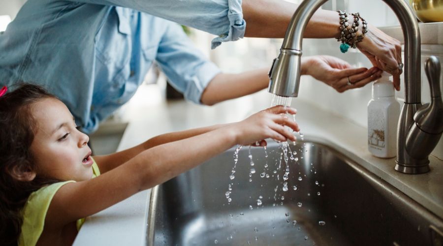 Ein kleines Mädchen mit dunklen Haaren und einem gelben Oberteil streckt seine Hände unter einen laufenden Wasserhahn in einem Küchenwaschbecken. Die Hände einer erwachsenen Person daneben betätigen einen Seifenspender. Das Bild stellt die alltägliche Nut