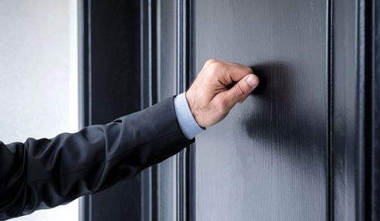 Eine männliche Hand im dunklen Anzug klopft an eine geschlossene, schwarz lackierte Holztür. Die Geste symbolisiert das Verlangen nach Einlass in eine Wohnung.