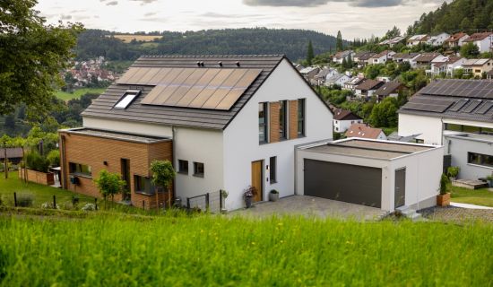 Modernes Einfamilienhaus mit weißen Wänden, Holzverkleidung und Solarpaneelen auf dem Dach, umgeben von grünem Gras und weiteren Häusern in hügeliger Landschaft unter bewölktem Himmel.