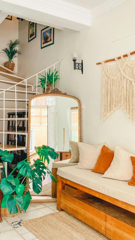 Eine große, üppige Monstera-Pflanze mit tief eingeschnittenen, leuchtend grünen Blättern steht in einem geflochtenen Korb auf dem Boden. Im Hintergrund ist auf der Stufe einer weißen Treppe eine weitere, kleinere Grünpflanze mit schmalen, spitzen Blättern