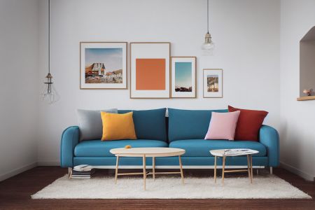 Ein tiefblaues Sofa steht vor einer schlichten weißen Wand, an der vier minimalistische Bilderrahmen hängen. Auf dem Sofa bilden Kissen in Senfgelb, Rosa und Rostrot einen lebendigen Kontrast zur kühlen Farbe des Polsters.