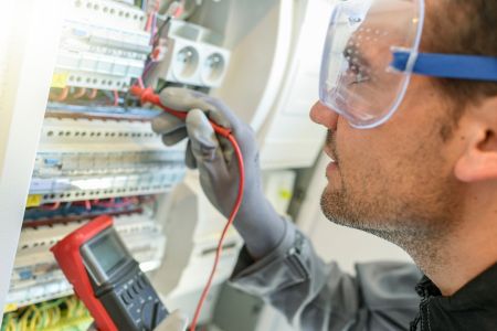 Ein Elektriker mit Schutzbrille und Arbeitshandschuhen prüft die Anschlüsse in einem geöffneten Sicherungskasten. Er hält die Messspitzen eines Multimeters an die Installation, um die Spannung zu kontrollieren.