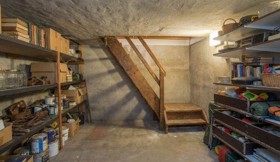 Ein älterer, vollgestellter Kellerraum mit grauen Betonwänden, niedriger Decke und einer einfachen Holztreppe. Regale an den Wänden sind dicht gefüllt mit Büchern, Dosen und Kisten; auf dem Boden stehen Farbeimer und Werkzeuge.