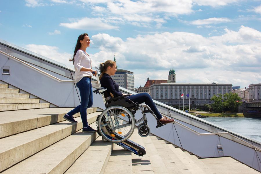 Junge Frau fährt mit einer anderen Frau im Rollstuhl einen großen Außentreppenaufgang mit Treppenraupe hinunter, im Hintergrund eine Stadtansicht.
