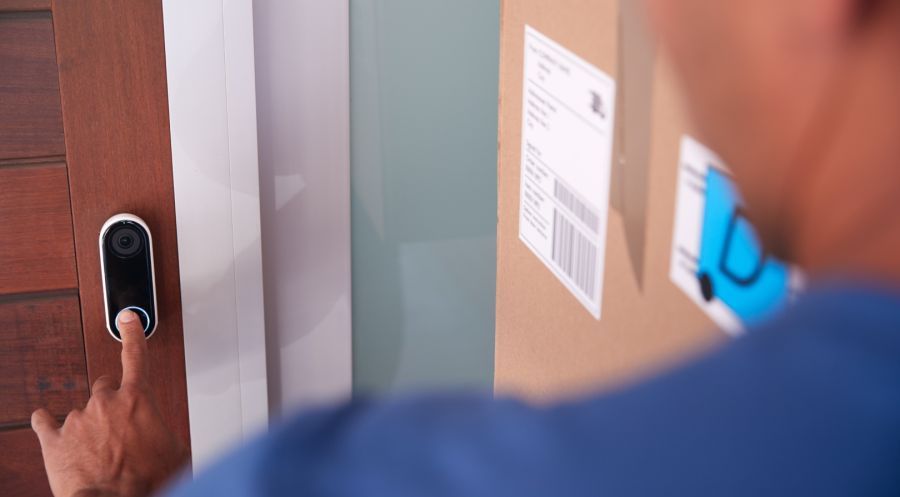 Nahaufnahme einer modernen Video-Türklingel an einer Holztür, die von einer Person in blauer Kleidung betätigt wird. Die Person trägt ein großes Paket unter dem Arm, auf dem Versandaufkleber sichtbar sind.