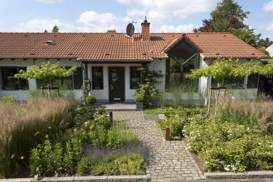 Bungalow, auf den ein leicht schräger, geklinkerter Weg zuführt durch den üppig bepflanzten Vorgarten