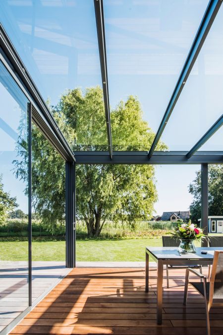 Innenansicht einer modernen Glas-Terrassenüberdachung mit schwarzem Metallrahmen. Die Decke besteht aus Glas, durch das der blaue Himmel und grüne Baumkronen zu sehen sind. Ein Holzboden und ein Esstisch mit Blumenarrangement sorgen für eine wohnliche Atm