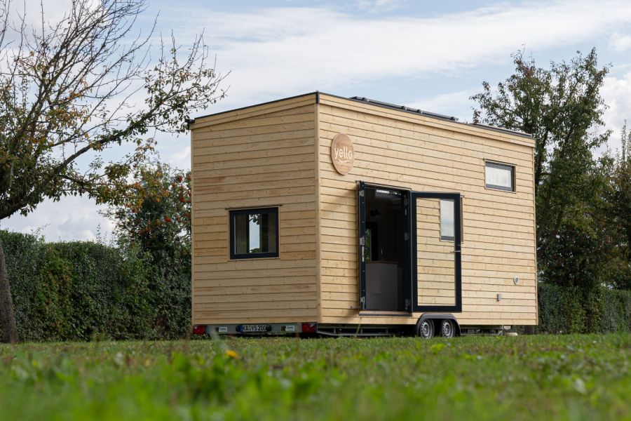 Ein Tiny House mit heller Holzfassade steht auf einem Anhänger (Trailer) auf einer grünen Wiese. Die Tür des Hauses steht offen und gibt den Blick auf eine kleine Küchenzeile frei.