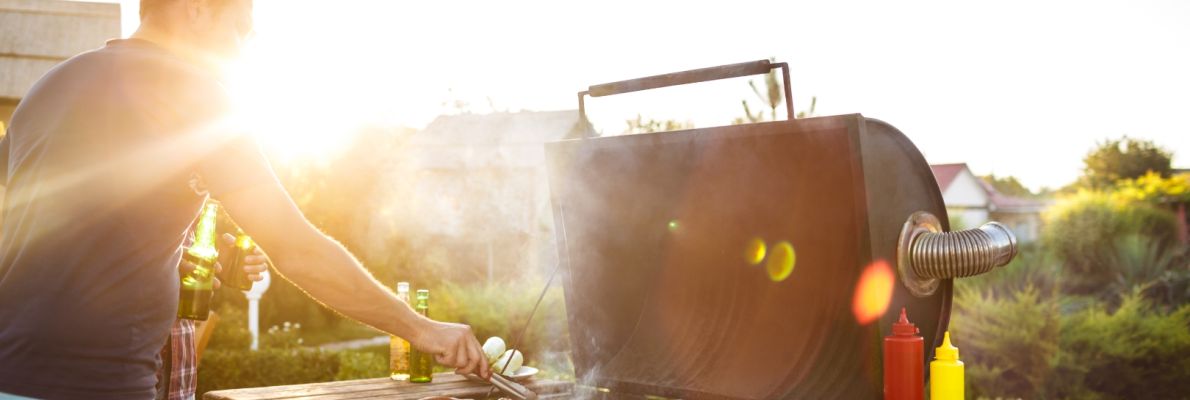 Ein Mann steht an einem großen Barbecue-Grill im Freien, während die warme Abendsonne im Hintergrund scheint. Er wendet Fleisch auf dem Rost mit einer Zange und hält in der anderen Hand eine Bierflasche. Auf einem Holztisch daneben stehen Burgerbrötchen,