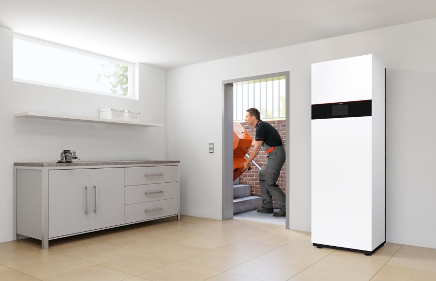 Ein Installateur trägt das orangefarbene Gehäusebauteil einer alten Heizanlage aus einem sauberen, hellen Kellerraum nach draußen. Rechts im Bild ist bereits die moderne, weiße Inneneinheit der neuen Wärmepumpe installiert.