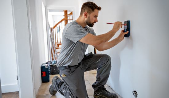 Ein Mann kniet auf dem Boden und markiert mit einem roten Stift durch die zentrale Öffnung eines Ortungsgeräts eine Stelle an der weißen Wand. Das Gerät liegt dabei ruhig an der Wand an, um die exakte Position zu bestimmen.