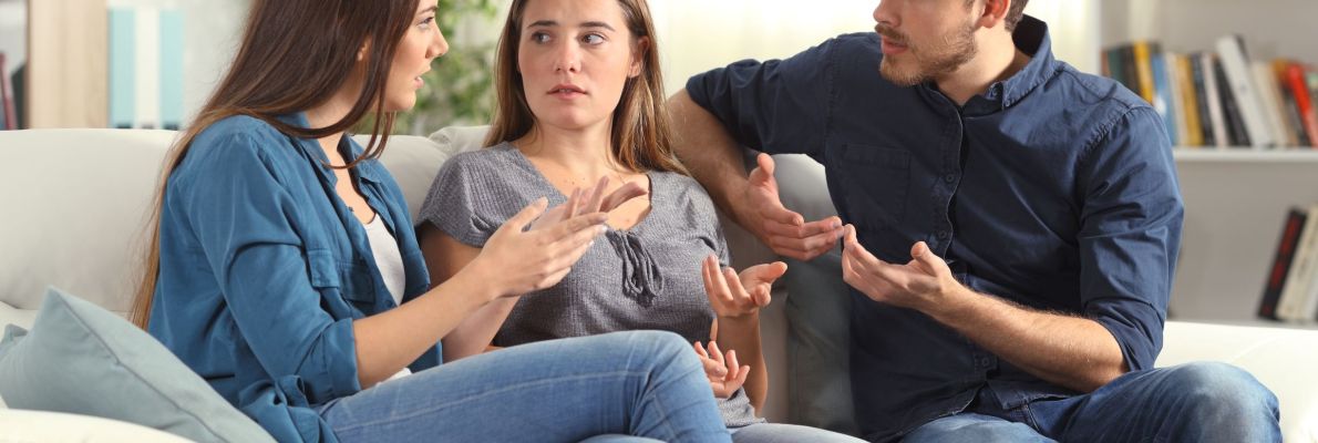 Drei junge Erwachsene sitzen nebeneinander auf einem hellen Sofa in einem Wohnzimmer. Eine Frau links und ein Mann rechts gestikulieren eindringlich mit den Händen und sprechen auf die in der Mitte sitzende Frau ein. Alle Beteiligten wirken angespannt und