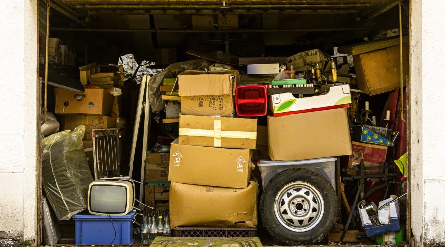 Ein überfülltes Garagentor zeigt eine chaotische Ansammlung von Kartons, Möbeln, einem alten Fernseher und verschiedenen Gegenständen, die den Raum nahezu vollständig ausfüllen. Die Szene vermittelt den Eindruck von Unordnung und Vernachlässigung.