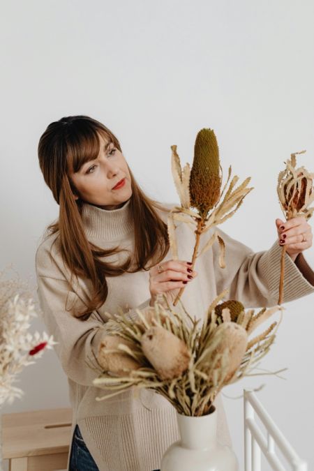 Eine Frau mit langen braunen Haaren und einem beigen Rollkragenpullover konzentriert sich darauf, große, getrocknete Banksia-Blüten in einer schlichten weißen Vase anzuordnen.