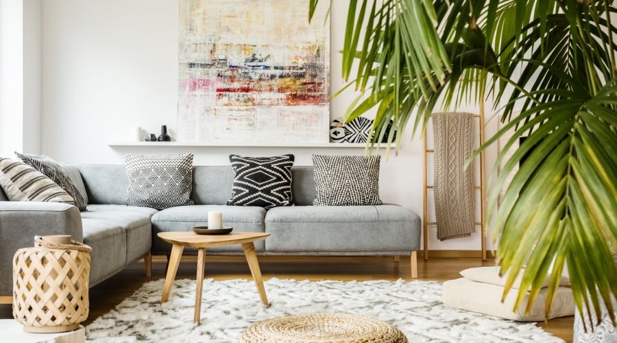 Ein helles, modernes Wohnzimmer mit einem großen grauen Ecksofa und verschiedenen schwarz-weiß gemusterten Kissen. Ein runder Jute-Pouf liegt auf einem hellen Teppich vor einem niedrigen Holztisch, während eine große Palme rechts den Raum mit Grün belebt.