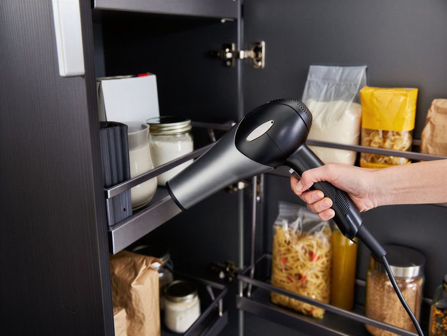 Eine Person hält einen modernen schwarzen Haartrockner in einen dunklen Vorratsschrank und richtet den Luftstrom gezielt auf die Ecken und Ritzen zwischen den Regalböden, in denen Vorräte wie Nudeln und Gläser stehen.
