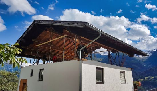 Ein komplettes Satteldach schwebt auf hydraulischen Stützen deutlich über dem gemauerten Erdgeschoss eines weißen Hauses; in der Lücke sind Bauarbeiter und Stützkonstruktionen vor einer alpinen Bergkulisse zu sehen.