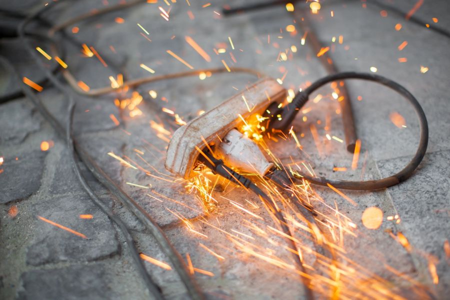 Eine alte, weiße Mehrfachsteckdose liegt auf dem Boden, in der zwei schwarze Stecker eingesteckt sind. Aus der Steckdose sprühen helle, glühende Funken in alle Richtungen, was auf einen Kurzschluss hindeutet.