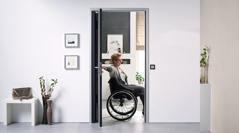 Ältere Frau fährt mit dem Rollstuhl aus dem Badezimmer, dabei hält sie die Tür auf