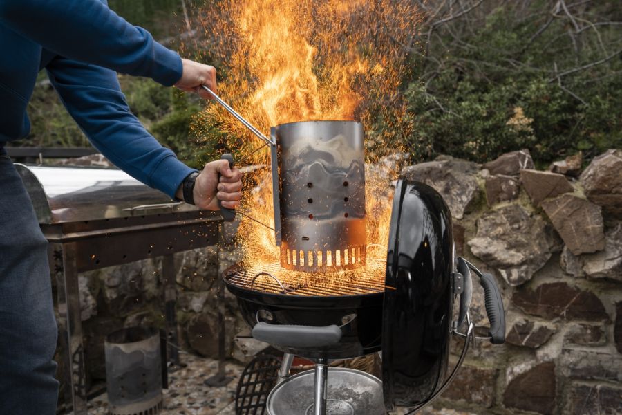 Eine Person hält einen Anzündkamin über einen runden Kugelgrill, aus dem hohe, helle Flammen und Funken herausschlagen. Der Mann trägt einen blauen Pullover und hält den heißen Behälter sicher am Griff.