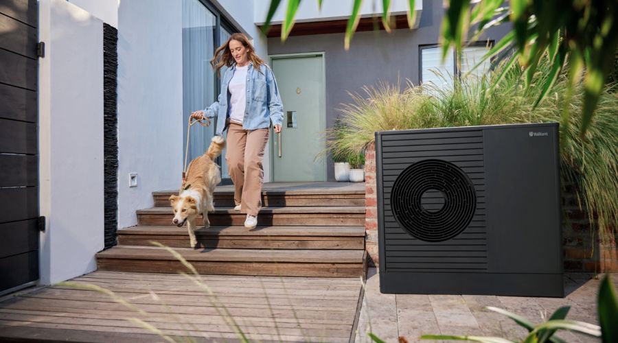 Eine Frau geht mit ihrem Hund eine flache Holztreppe vor einem modernen Einfamilienhaus hinunter. Rechts im Vordergrund steht eine kompakte, dunkelgraue Luft-Wasser-Wärmepumpe unauffällig im Außenbereich.