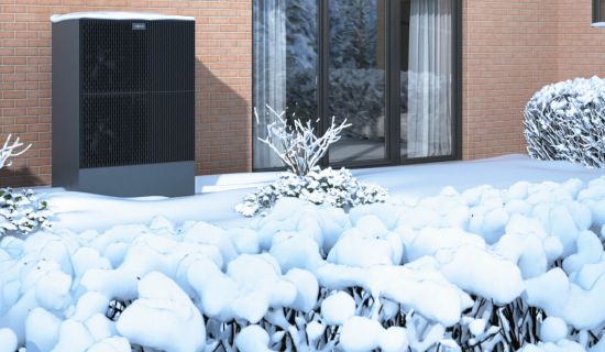 Eine moderne, dunkle Wärmepumpen-Außeneinheit steht neben einer Ziegelwand im verschneiten Garten vor einer großen Fensterfront. Die umliegenden Sträucher sind tief verschneit.