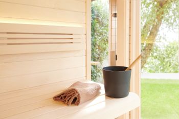 Heller Innenraum einer Holzsauna mit Saunaeimer, Handtuch und großem Fenster mit Blick ins Grüne.