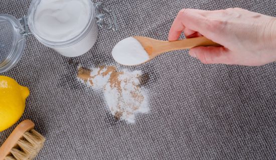 Eine Hand mit einem Holzlöffel streut weißes Natronpulver auf einen braunen Fleck auf einem grauen Teppich. Im Hintergrund sind ein Glasbehälter mit Natron, eine halbe Zitrone und eine kleine Bürste zu sehen. Die Szene illustriert die Verwendung von Hausm