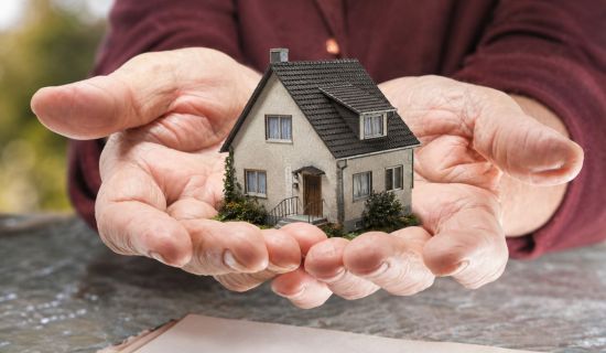 Die gealterten Hände einer Person halten schützend ein detailliertes Miniaturmodell eines Einfamilienhauses über einem offiziellen Dokument, auf dem das Wort „Testament“ zu lesen ist.