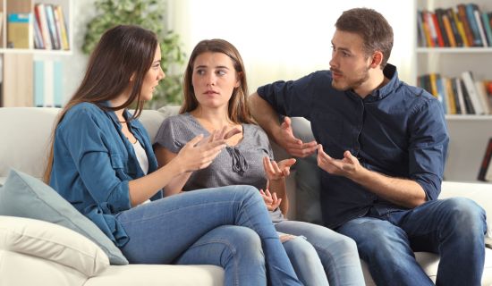 Drei junge Erwachsene sitzen nebeneinander auf einem hellen Sofa in einem Wohnzimmer. Eine Frau links und ein Mann rechts gestikulieren eindringlich mit den Händen und sprechen auf die in der Mitte sitzende Frau ein. Alle Beteiligten wirken angespannt und