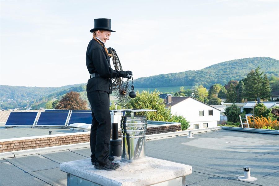 Eine Schornsteinfegerin steht auf einem Flachdach und reinigt einen Schornstein. Sie trägt die traditionelle schwarze Schornsteinfegeruniform mit Zylinderhut und Handschuhen. In der Hand hält sie eine Reinigungsbürste mit Kugelgewicht an einer Kette. Im H