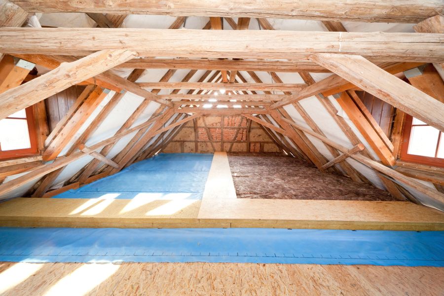 Ein heller Dachboden im Rohbau mit freiliegenden Holzbalken, auf dessen Fußboden blaue Dampfbremsfolien und Dämmplatten aus Mineralwolle verlegt werden.