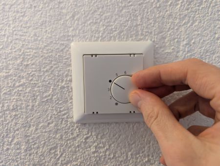 Eine Hand dreht an einem Thermostat-Drehregler an einer Wand. Der Regler zeigt Zahlen von 0 bis 6 und befindet sich auf einer strukturierten Wandoberfläche.