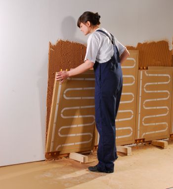 Frau legt Klimaplatten mit wasserführenden Rohren auf einer Holzweichfaserplatte, mit Lehm verputzt, an eine Wand an