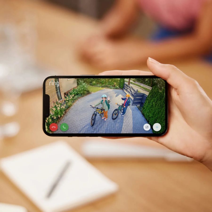 Eine Hand hält ein Smartphone, auf dessen Bildschirm die Live-Ansicht einer gepflasterten Hauseinfahrt zu sehen ist. Auf dem Display sind zwei Personen mit Fahrrädern erkennbar, die in die Kamera winken.