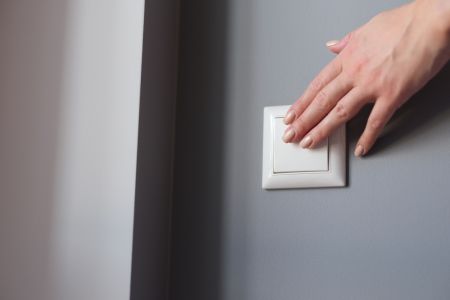 Eine weibliche Hand mit lackierten Fingernägeln betätigt einen schlichten, weißen Lichtschalter. Der Schalter ist auf einer mattgrau gestrichenen Wand montiert, links im Bildausschnitt ist ein Teil eines weißen Türrahmens zu sehen.