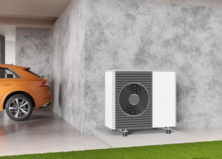 Die weiße Außeneinheit einer modernen Luft-Wasser-Wärmepumpe mit dunklen Lamellen und rundem Ventilatorgitter steht auf einem flachen Sockel vor einer grauen Betonwand. Links daneben parkt das Heck eines orangefarbenen Autos. Im Vordergrund grenzt ein grü