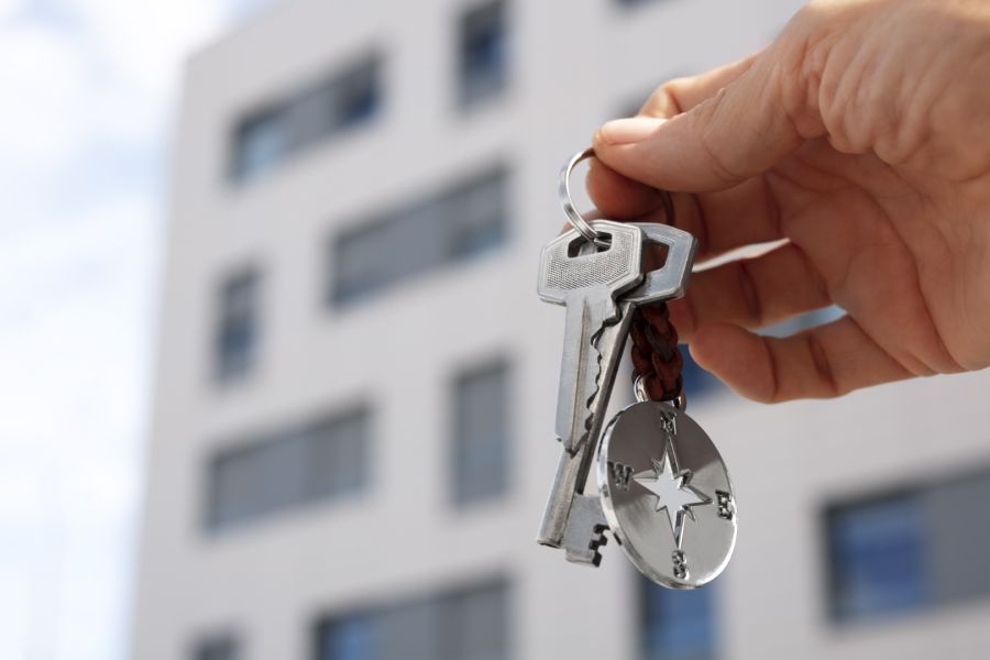 Eine Hand hält einen metallenen Schlüsselbund mit einem markanten Kompass-Anhänger hoch in die Luft. Im unscharfen Hintergrund ist die helle Fassade eines modernen Mehrfamilienhauses zu erkennen.