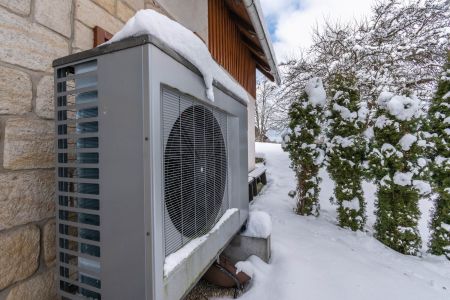 Nahaufnahme einer grauen Wärmepumpe an einer Natursteinwand im Winter. Auf dem Gehäuse liegt eine dicke Schneeschicht, während im Hintergrund verschneite Tujen zu sehen sind.