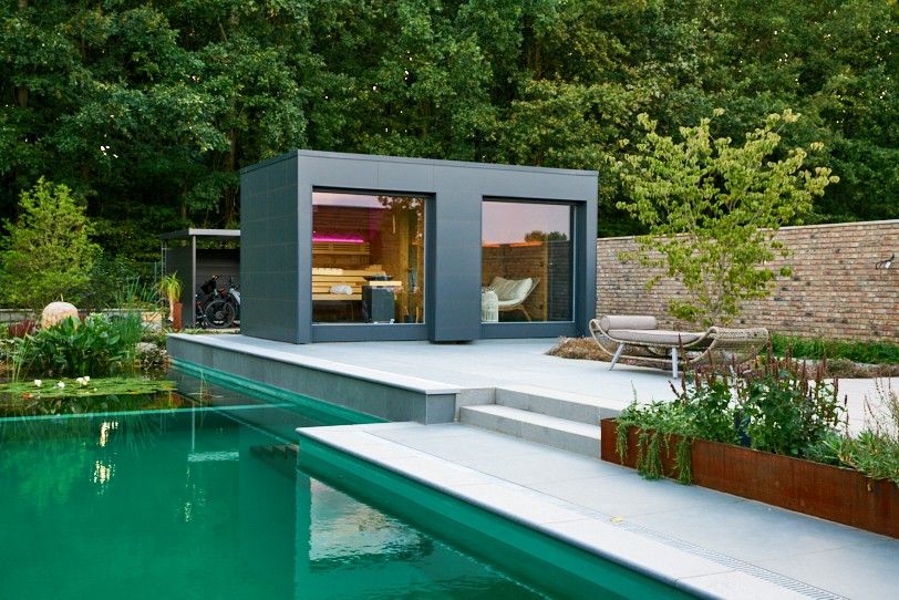 Modernes kubisches Saunahaus mit dunkler Fassade, zwei großen Fensterfronten und Terrasse an einem Wasserbecken.