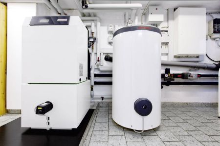 Eine bodenstehende, große Wärmepumpen-Inneneinheit in Weiß steht neben einem passenden, zylindrischen Warmwasserspeicher in einem typischen Heizungskeller. Im Hintergrund sind zahlreiche isolierte Rohrleitungen, Messgeräte und elektrische Steuereinheiten